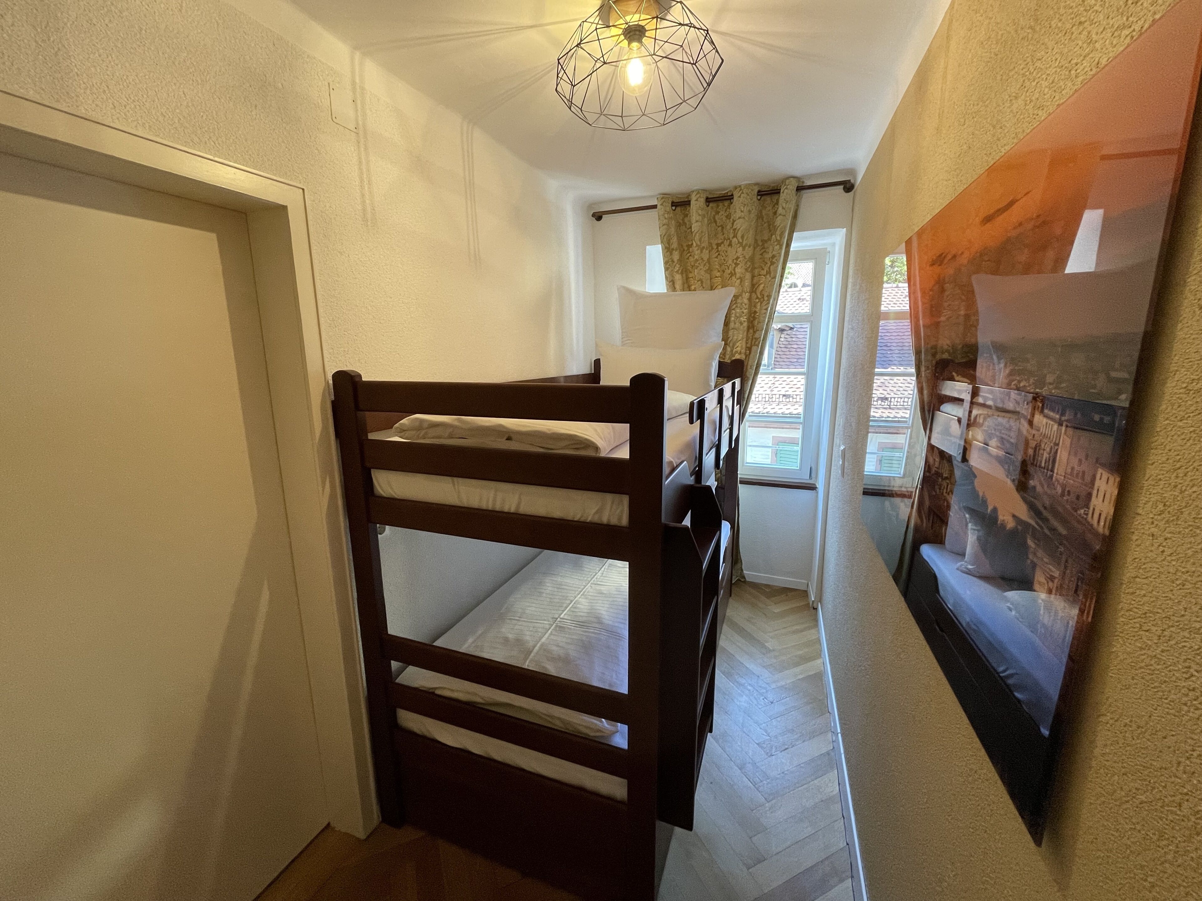A narrow hallway with a bunk bed, a ceiling light fixture, a window with curtains, and a wall-mounted picture.