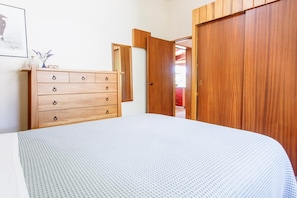 A bedroom with a wooden dresser, a bed with a white textured bedspread, and a wooden wardrobe. - 1970's Renovated, self contained comfortable 2 bedroom bach (Wanaka)