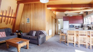 A cozy living room with a sofa, a coffee table, and a kitchen area with wooden cabinets and a dining table. - 1970's Renovated, self contained comfortable 2 bedroom bach (Wanaka)