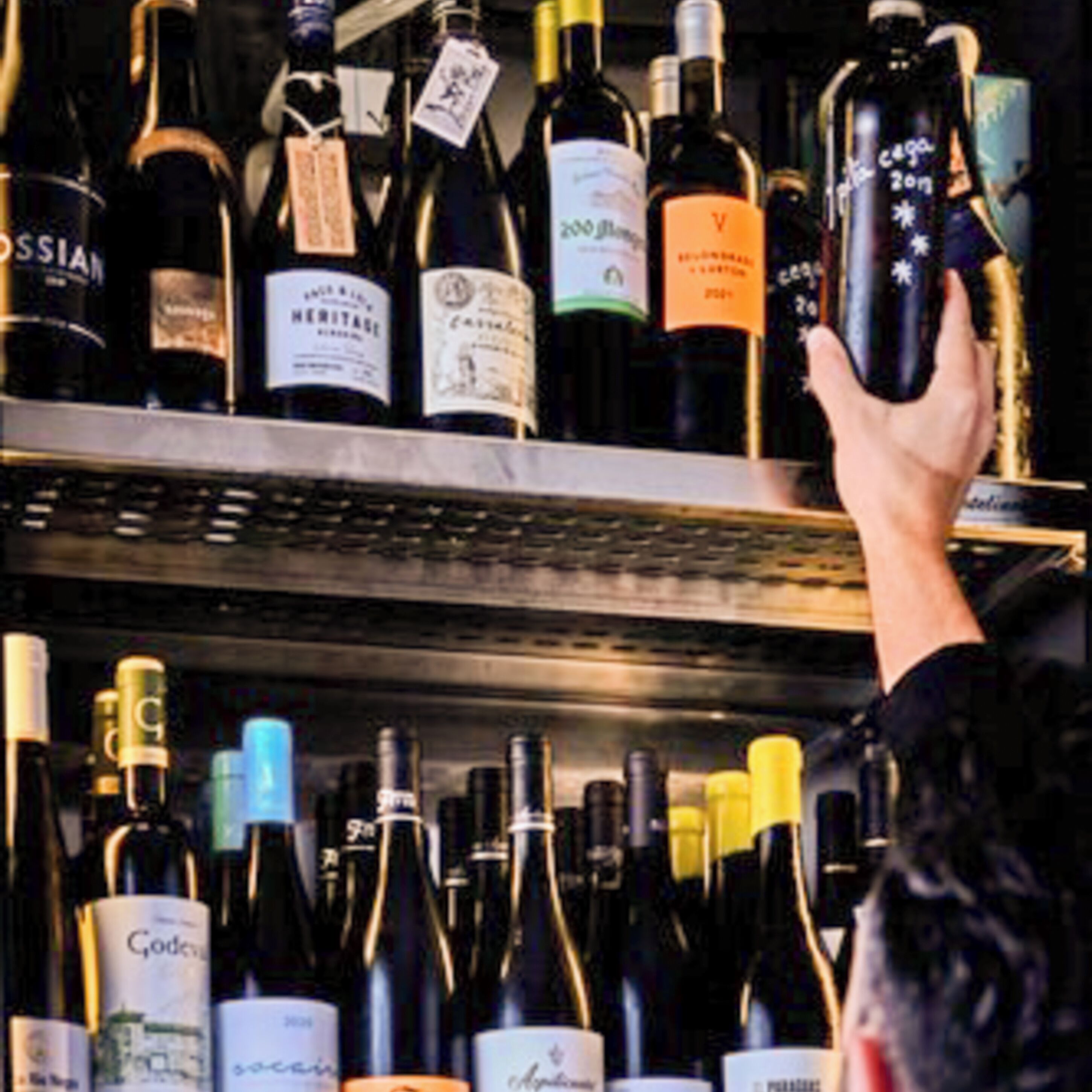 Una mano alcanza una botella en un estante lleno de diversas botellas de vino.