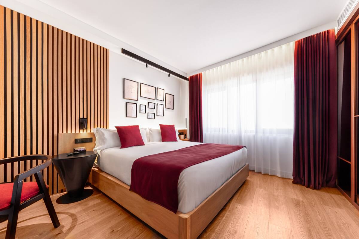 A modern hotel room with a wooden bed, red accents, and a large window with curtains.