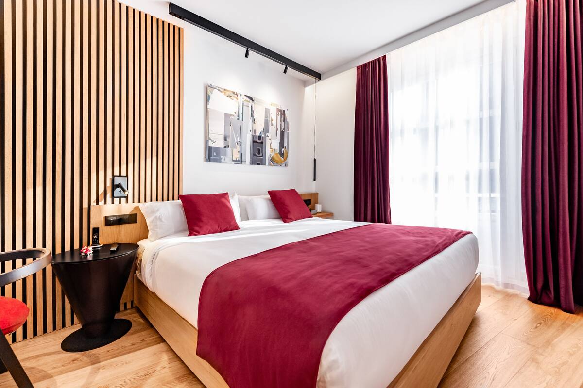 A modern hotel room with a large bed, red and white bedding, a wooden slat wall, a black side table, and a red chair.