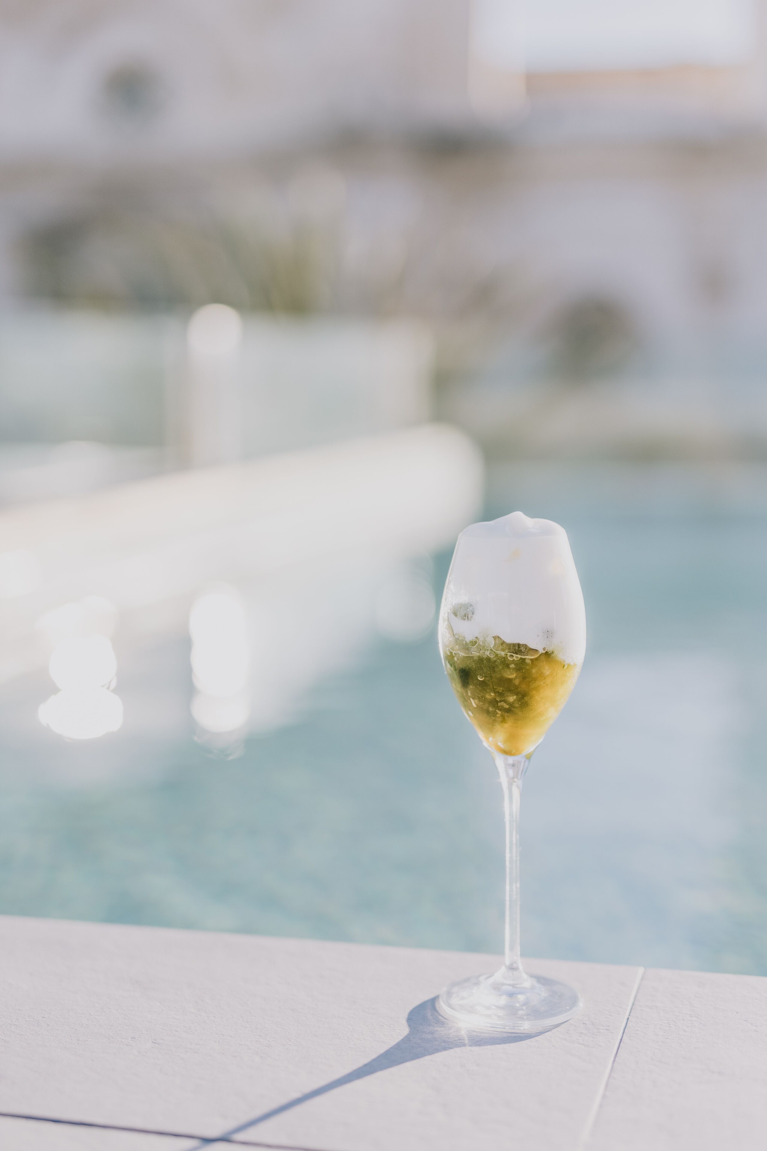 Una copa de vino blanco sobre una superficie blanca cerca de una piscina.