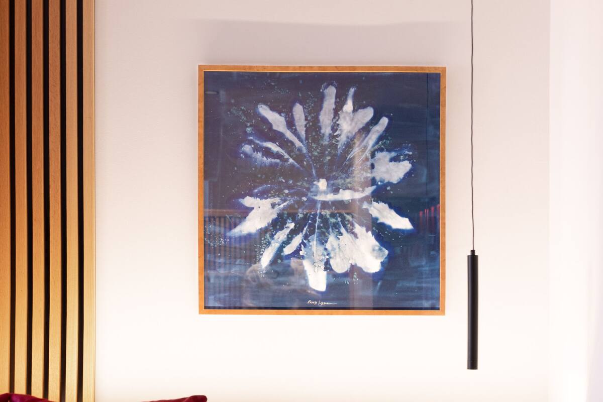 A modern hotel room with a wooden headboard, a red pillow, a white bedspread, a digital clock, and a framed abstract artwork on the wall.