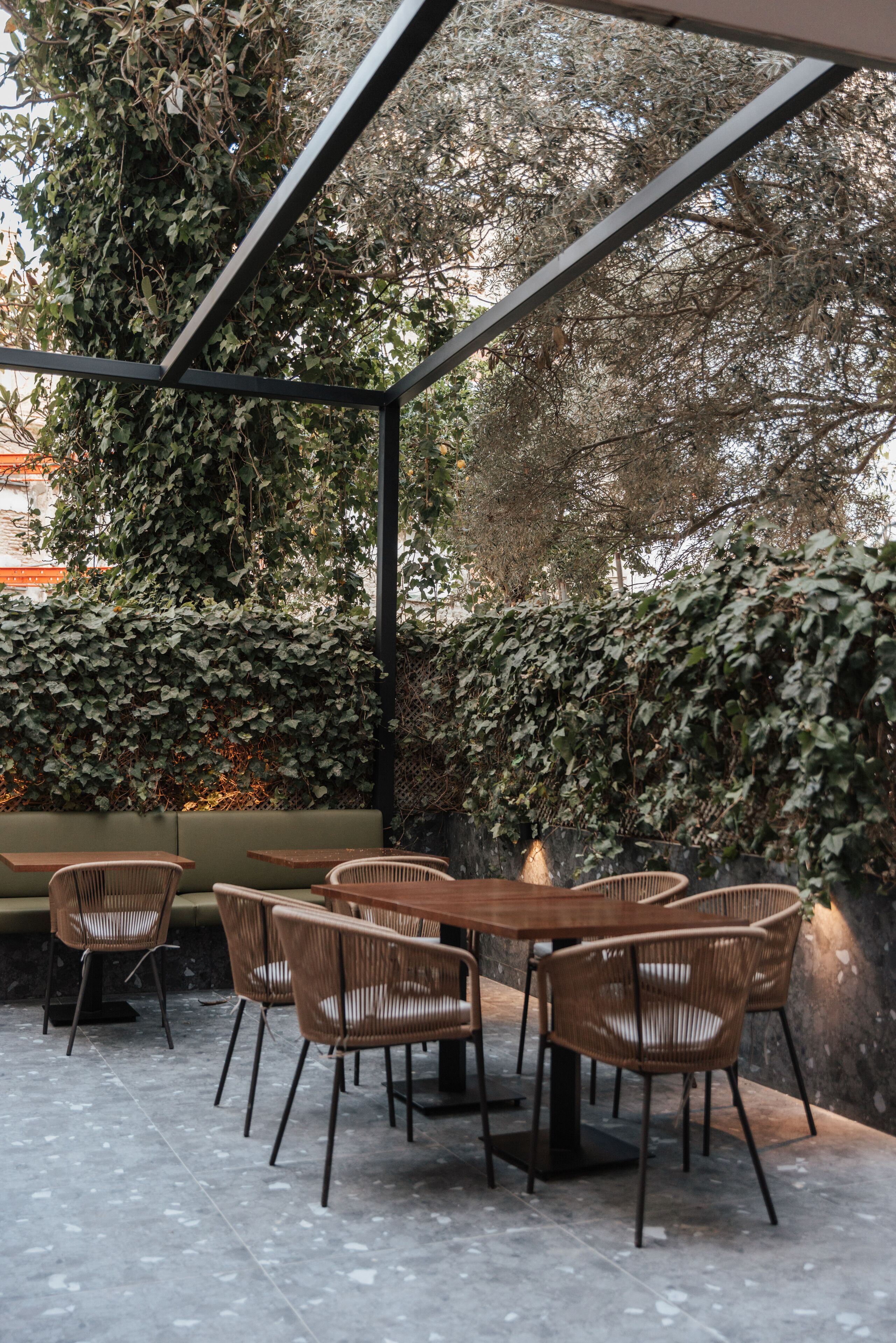 Un área de estar al aire libre con sillas de mimbre y una mesa de madera, rodeada de abundante vegetación y una estructura de metal.