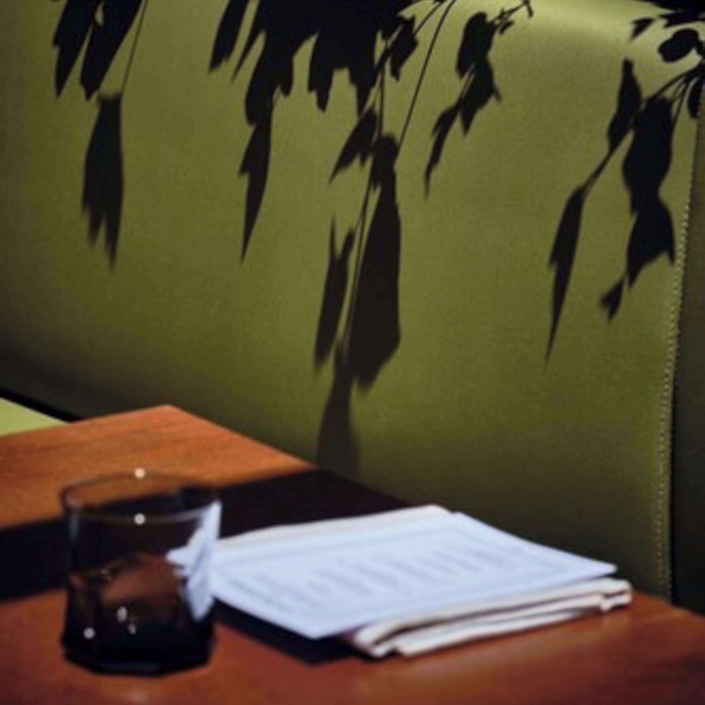 Una silla tapizada de color verde con estampado de hojas, una mesa de madera con un vaso y papeles, y un bolso oscuro.