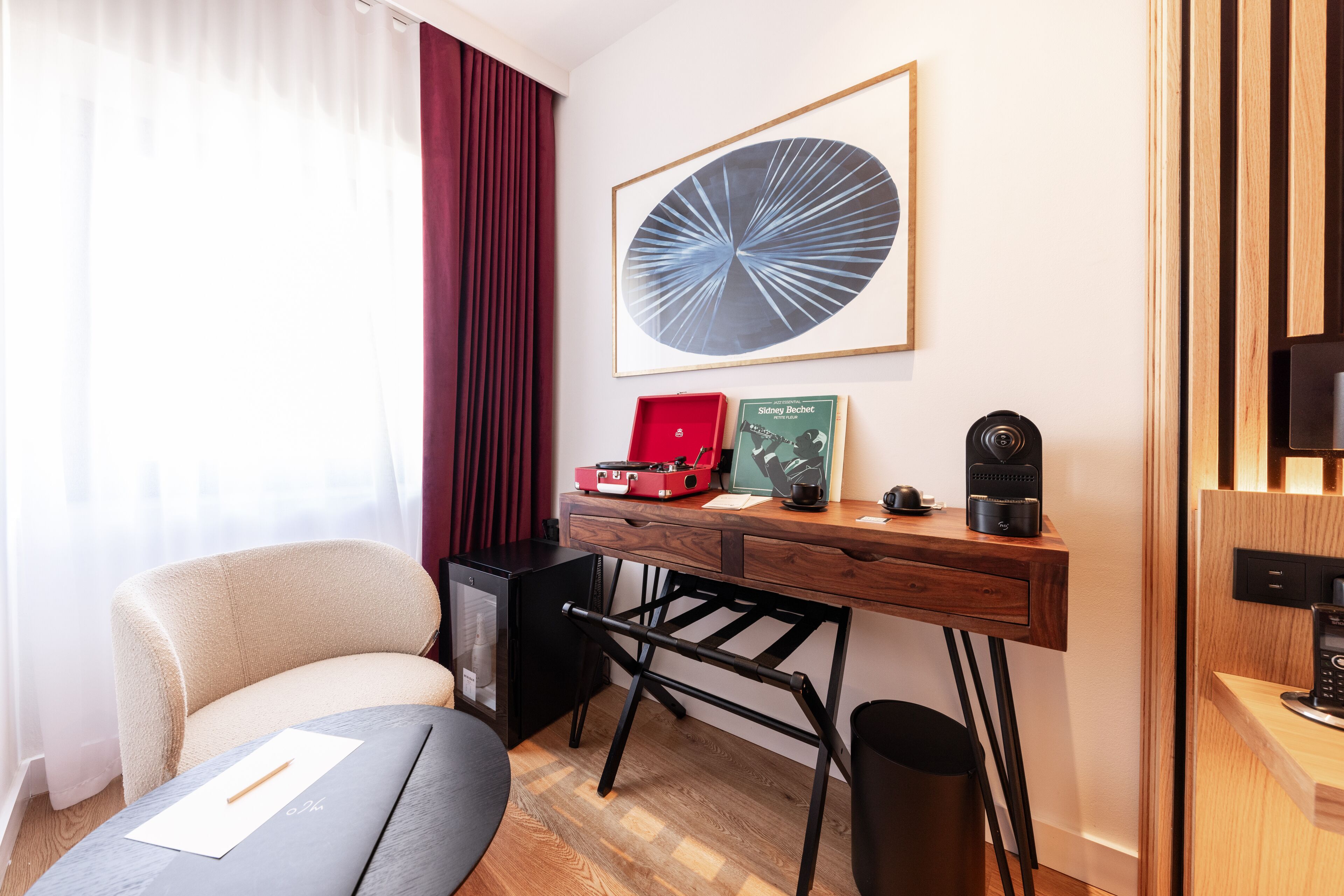 Una habitación de hotel con un escritorio de madera, una silla, un mueble negro pequeño, una maleta roja, un cuadro enmarcado y una máquina de café.