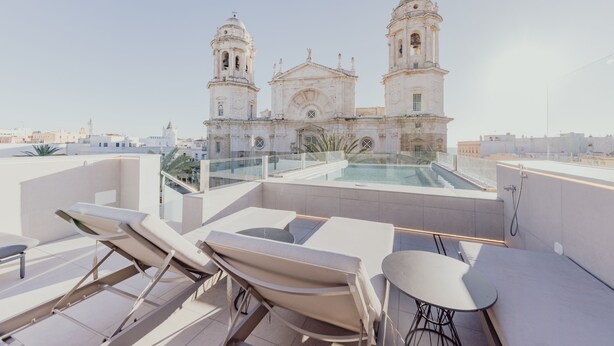 Terraza en la azotea con sillones de descanso y piscina, con vistas a un edificio histórico con dos campanarios.