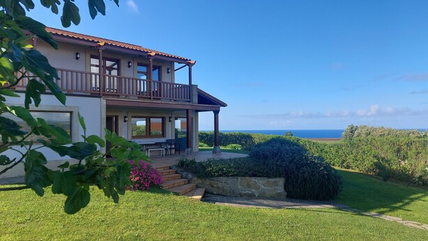 Una casa de dos pisos con balcón, un patio con mobiliario exterior y vista al océano.