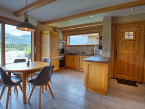 Une cuisine moderne avec des meubles en bois, une table à manger avec des chaises et une vue sur les montagnes grâce à de grandes fenêtres.