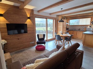 Een modern interieur met een aan een houten wand bevestigde televisie, een eethoek met stoelen, een keuken met houten kastjes en een grote schuifpui met uitzicht.