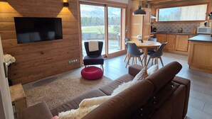 A modern interior with a wooden wall-mounted TV, a dining area with chairs, a kitchen with wooden cabinets, and a large sliding glass door leading to a view. - Quiet 2-bedroom apartment with terrace in Samoëns (Samoëns)
