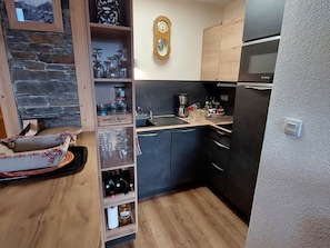 A compact kitchen with dark cabinets, a Bosch refrigerator, a wooden shelf with wine bottles, and a clock on the wall.