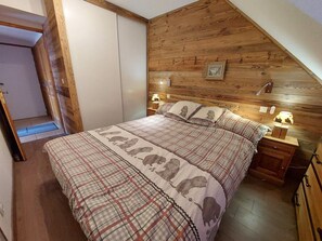 A bedroom with a wooden sloped ceiling, a bed with a plaid bedspread, two bedside lamps, and a small bedside table.