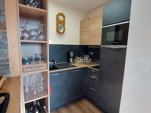 A compact kitchen with dark cabinets, a Bosch microwave, a wooden shelf with wine glasses, and a mirror on the wall.
