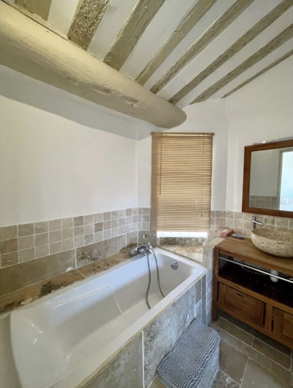 A bathroom with a freestanding bathtub, a wooden vanity with a large mirror, and a window with blinds.