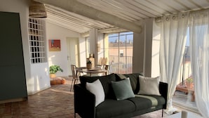 A cozy living room with a black sofa, a white table, and a patterned rug. There is a dining area with a table and chairs, and a balcony with potted plants. - Magnificent apartment with terrace and rooftop view (Aix-en-Provence)