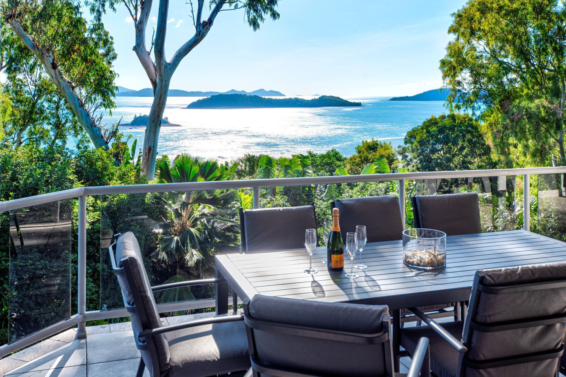 A table with four chairs, a bottle of champagne, and glasses on a balcony overlooking a body of water and greenery.