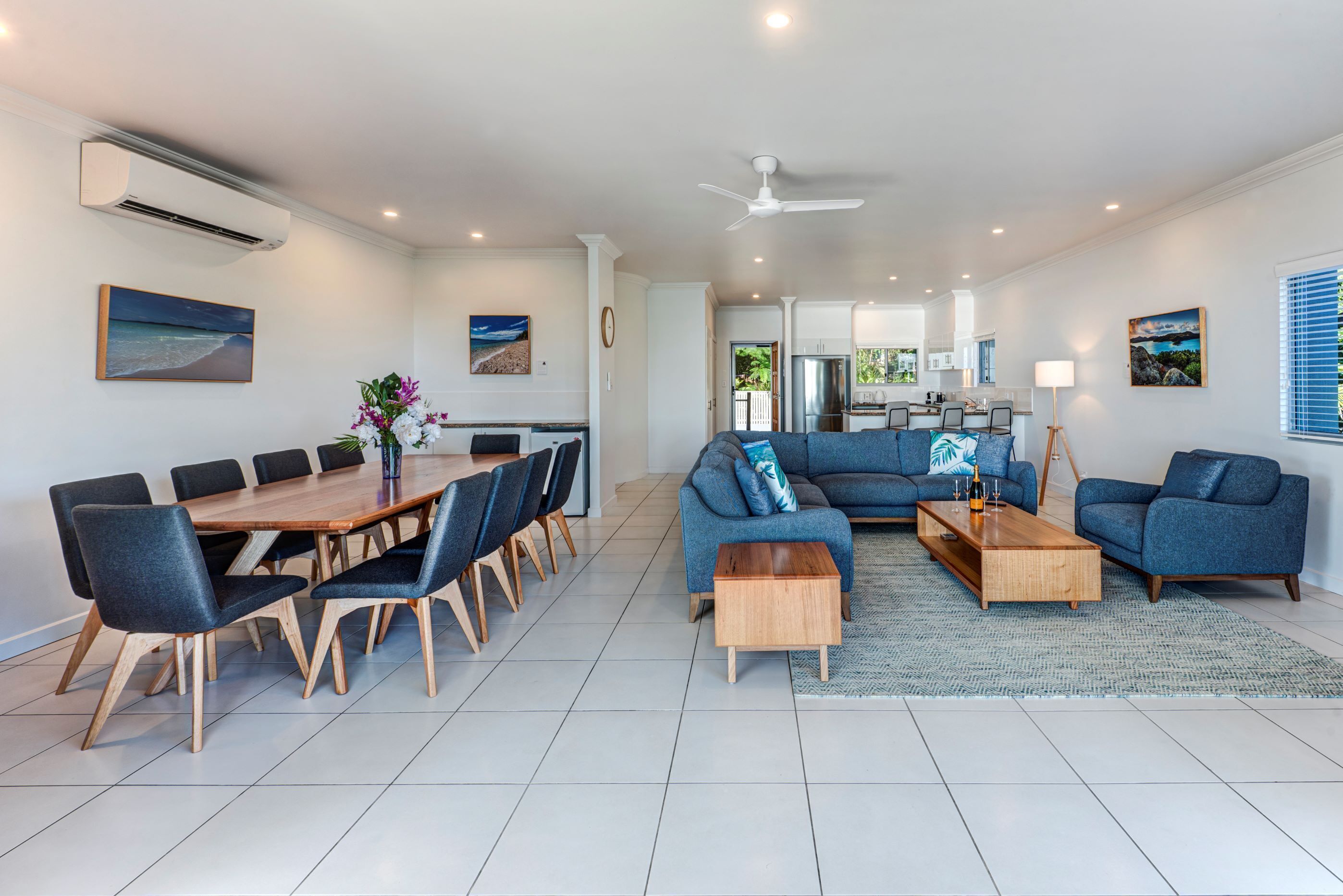 A modern living room with a dining area, blue sofas, a wooden coffee table, and a large window.