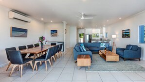 A modern living room with a dining area, blue sofas, a wooden coffee table, and a large window.