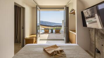 Habitación de hotel con cama, televisión y vistas a un patio con mobiliario exterior y una montaña al fondo.