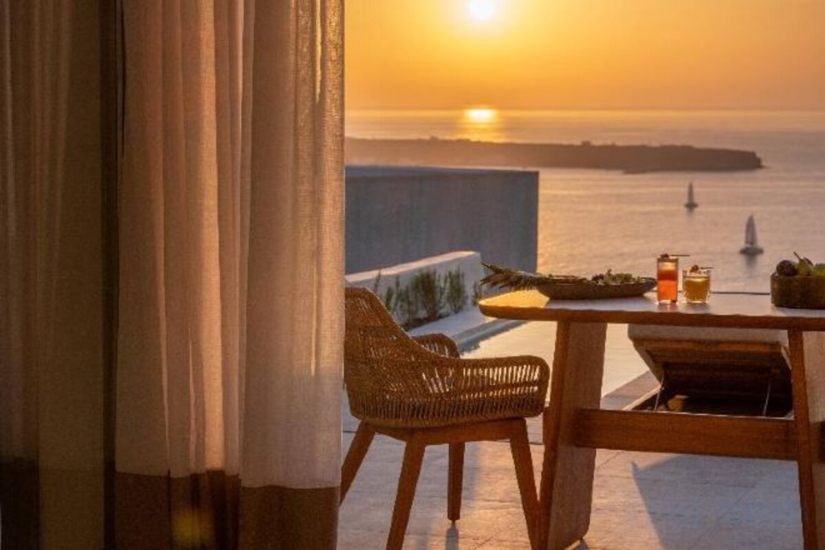 A balcony with a view of the sea during sunset, a wicker chair, a table with a bottle and a glass, and a small plant.