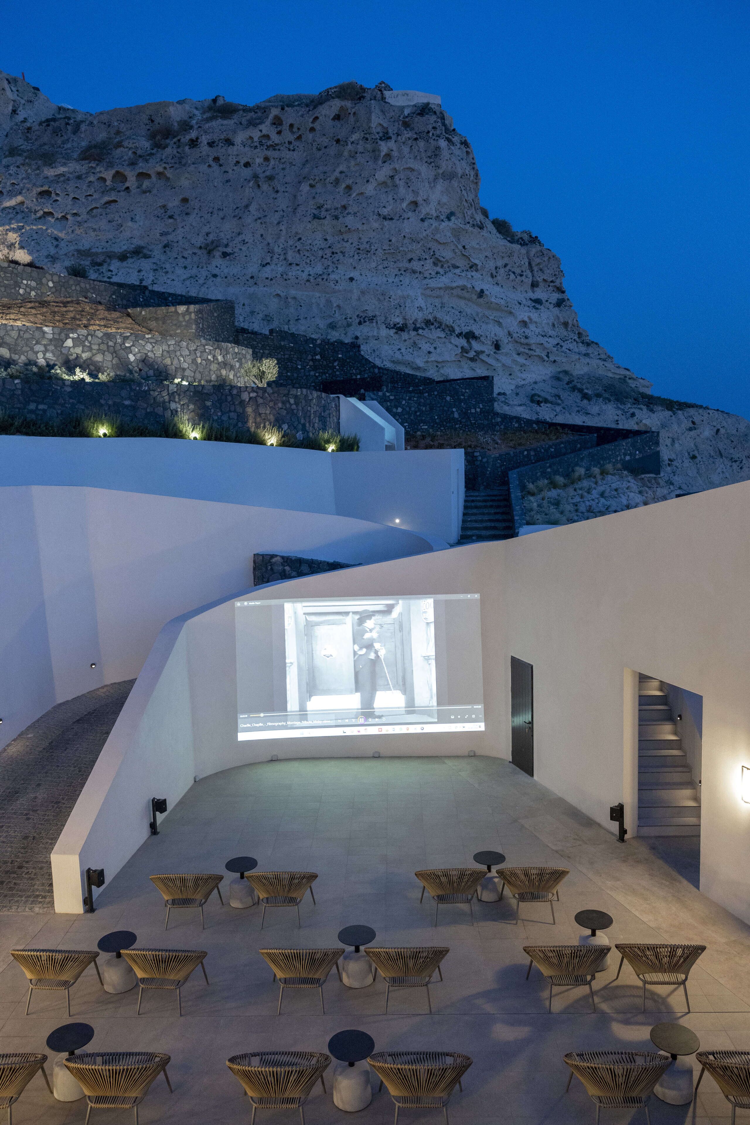 Zona d'estar a l'aire lliure amb cadires i taules de vimetla, una pantalla gran que mostra una vista interior i una escala que puja a un edifici amb un fons rocos.