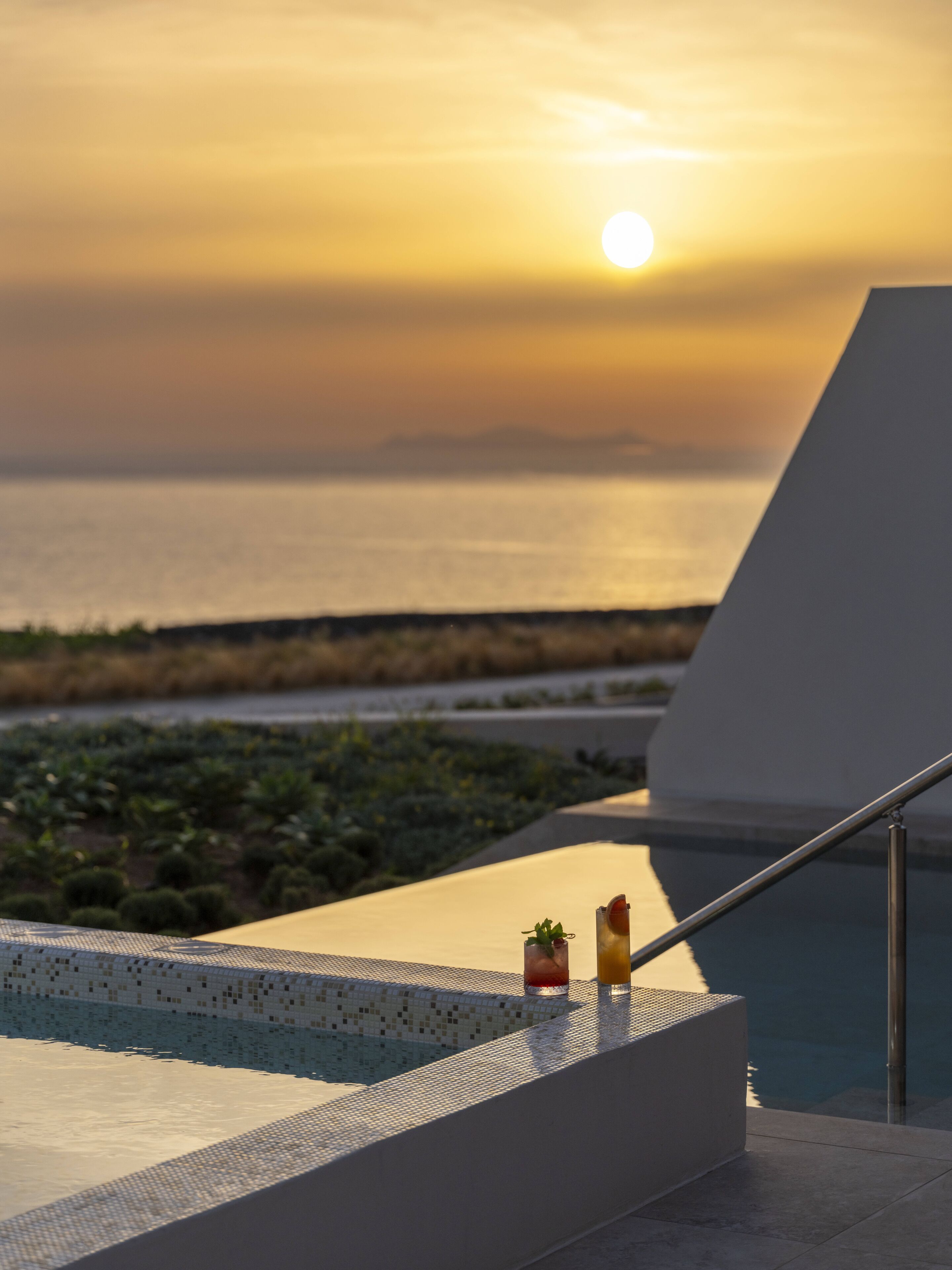 Vista à beira da piscina com o pôr do sol sobre o oceano, duas bebidas na borda e um design arquitetônico moderno.