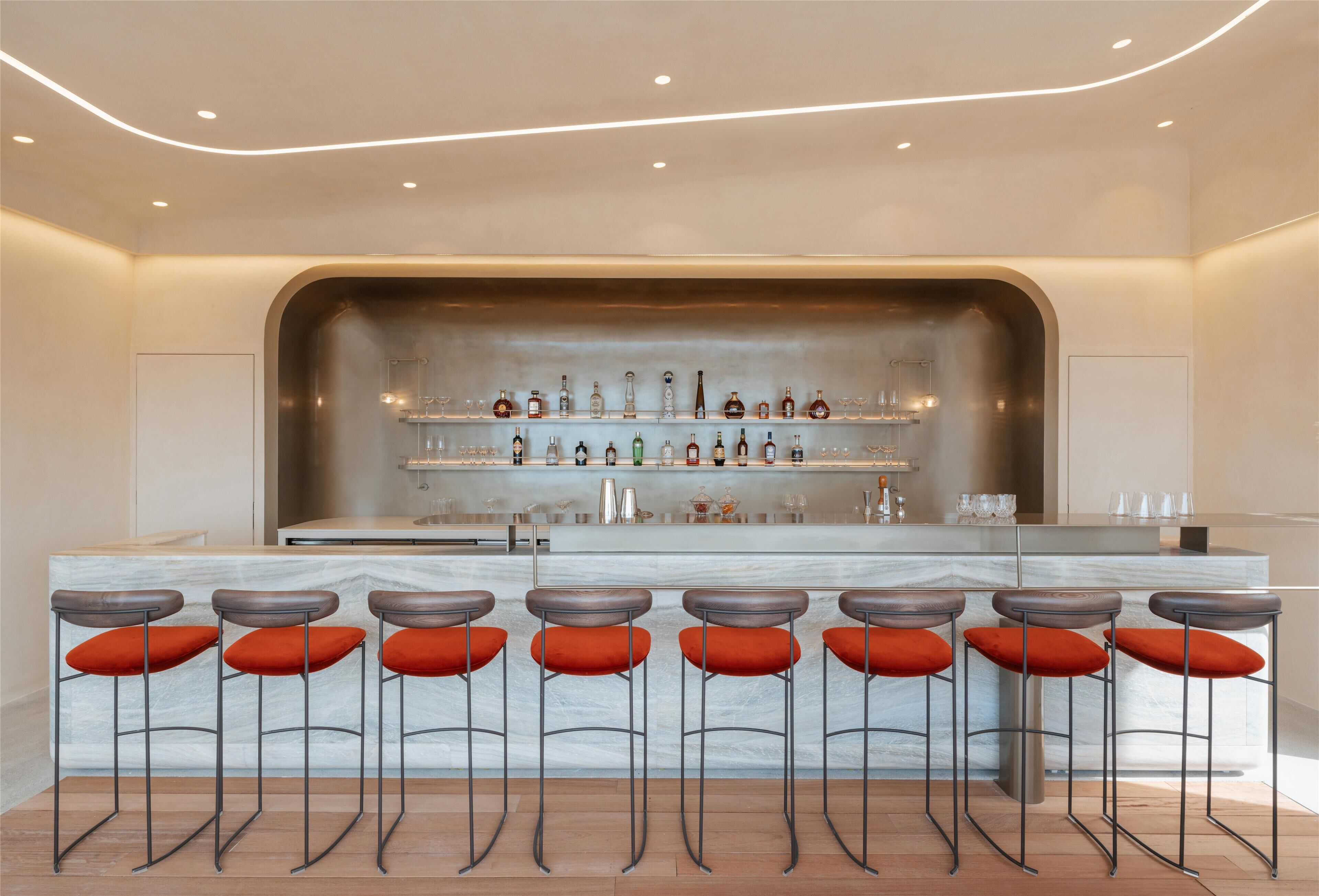 A modern bar area with a marble countertop, multiple stools with orange cushions, and a variety of bottles and glasses on shelves.