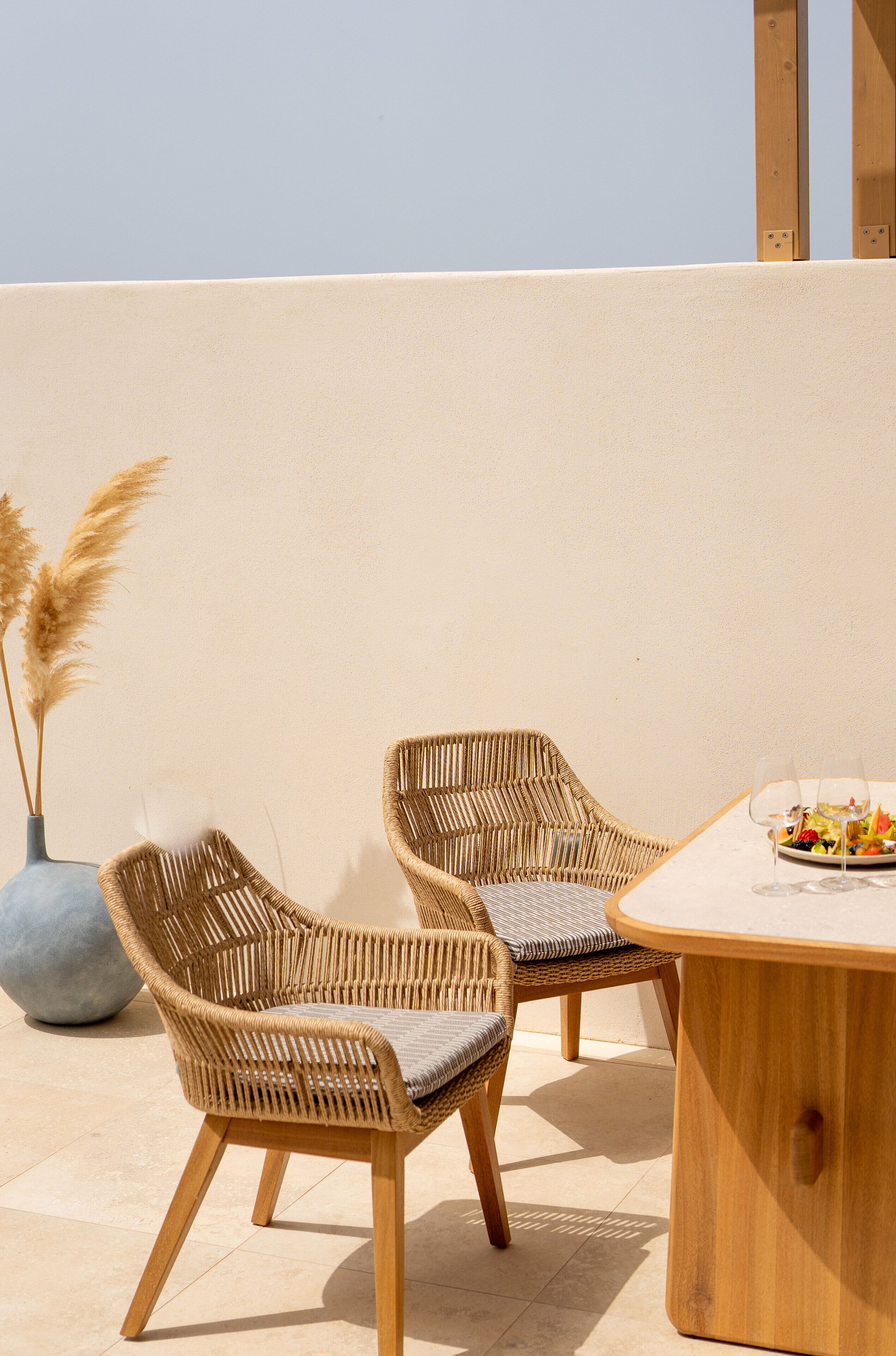 Two wicker chairs with cushions, a round wooden table, and a vase with pampas grass.