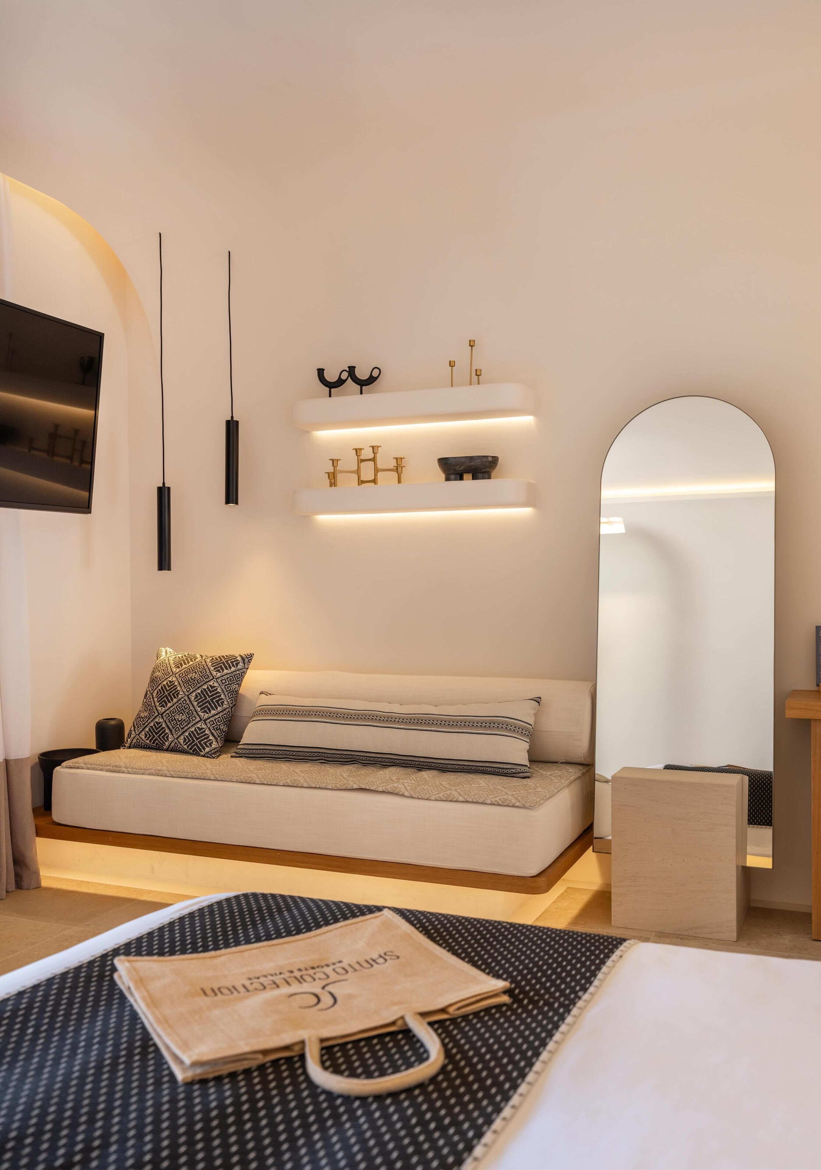 A modern hotel room with a sofa, a mirror, a flat-screen TV, and a small shelf with decorative items.