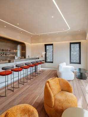 A modern bar area with stools and a bar counter, a seating area with sofas, and large windows providing natural light.