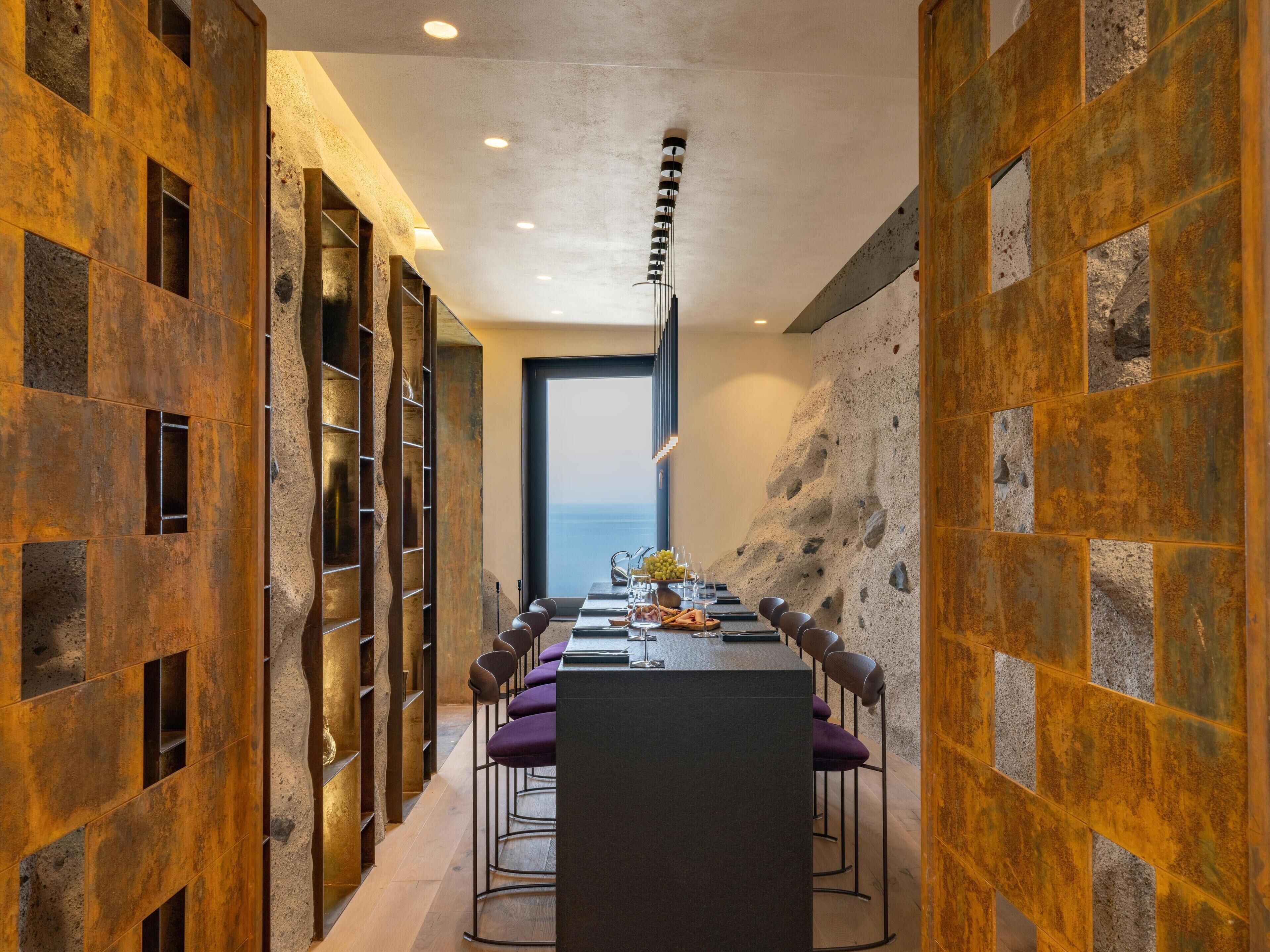 A modern dining area with a long table, purple chairs, and a view of the sea through a large window.