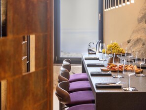 A modern dining area with a long table set for a meal, featuring a variety of cheeses, fruits, and glasses.