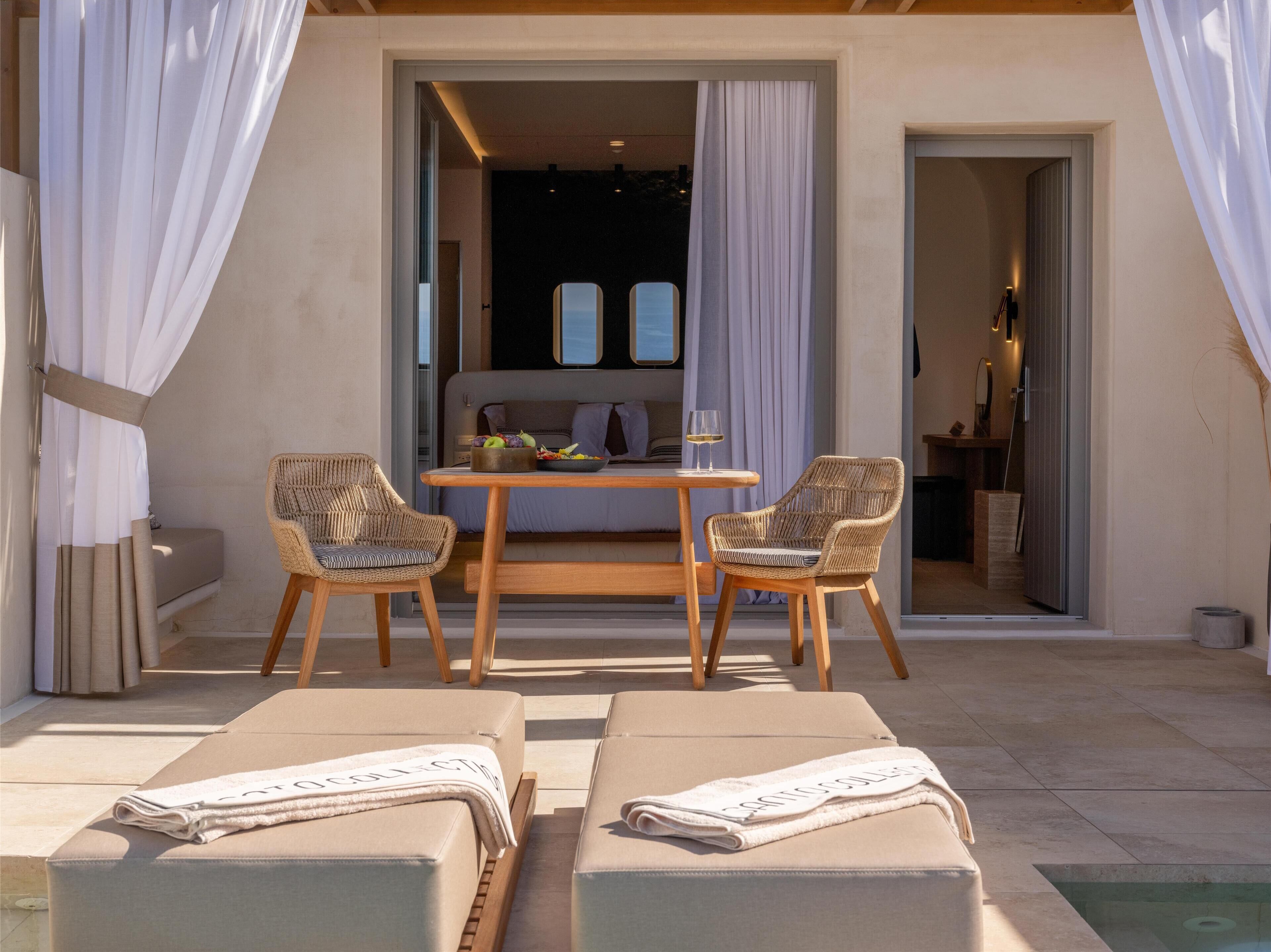 A sunlit outdoor seating area with two wicker chairs and a wooden table, a sofa, and two lounge chairs with towels.