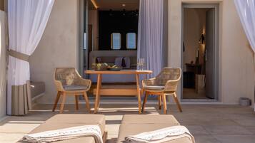 A sunlit outdoor seating area with two wicker chairs and a wooden table, a sofa, and two lounge chairs with towels.