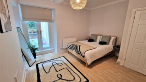 A bedroom with a bed, a hanging light fixture, a window with blinds, and a door. - Seafront Apartment with Garden - Perfect location in Mumbles (Swansea)