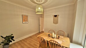 A dining area with a wooden table, chairs, a hanging light fixture, a wall-mounted macrame piece, and a framed map. - Seafront Apartment with Garden - Perfect location in Mumbles (Swansea)