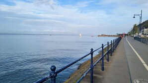 A coastal promenade with a metal railing, a paved walkway, and a clear view of the sea. - Seafront Apartment with Garden - Perfect location in Mumbles (Swansea)