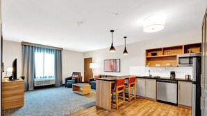 A modern hotel room with a kitchenette, a dining area with a bar, a living room with a sofa and a coffee table, and a large window with curtains. - Candlewood Suites Detroit - Auburn Hills by IHG (Auburn Hills)