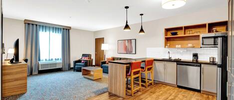 A modern hotel room with a kitchenette, a dining area with a bar, a living room with a sofa and a coffee table, and a large window with curtains.