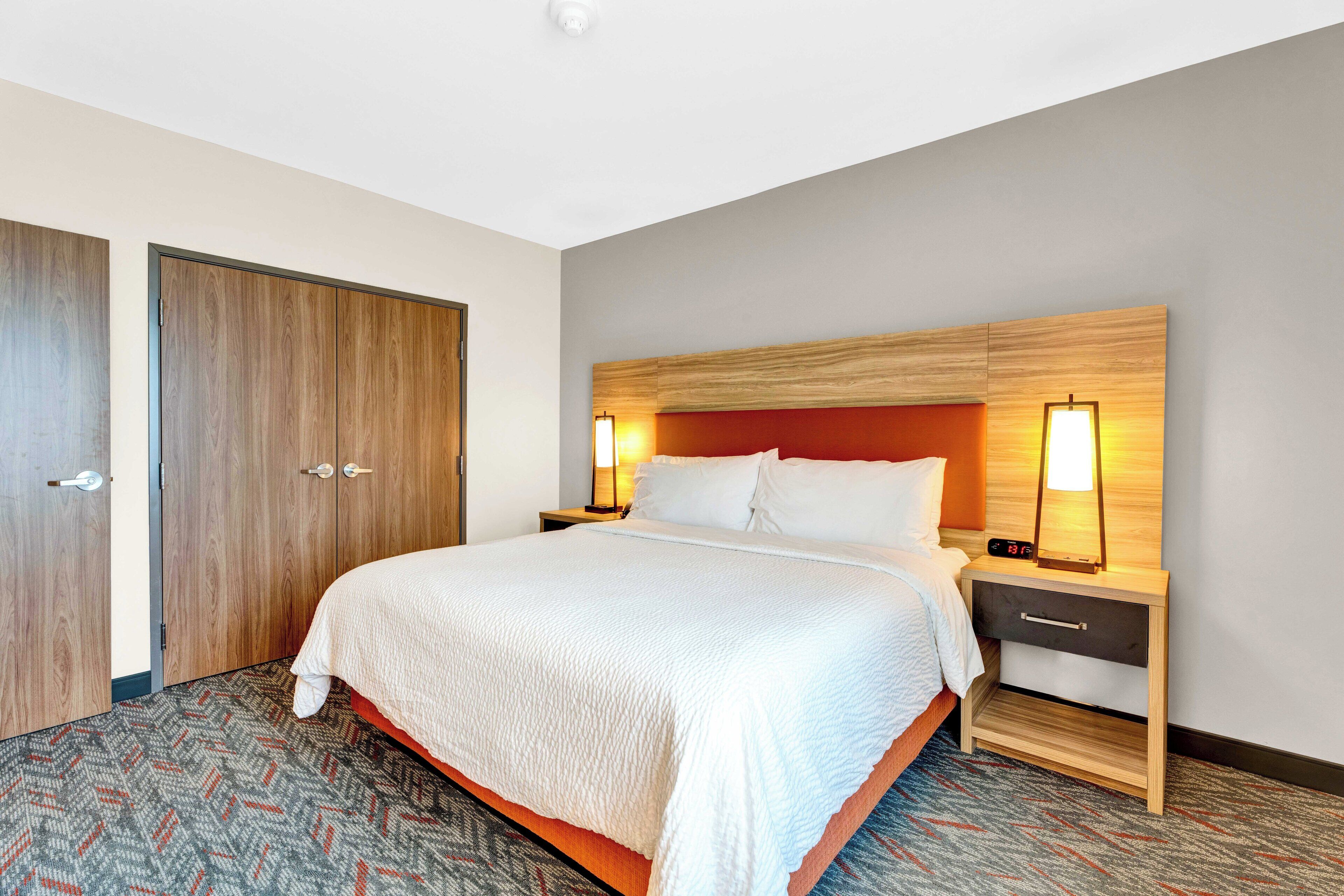 A hotel room with a large bed, two bedside tables, a nightstand, a wooden headboard, and a patterned carpet.
