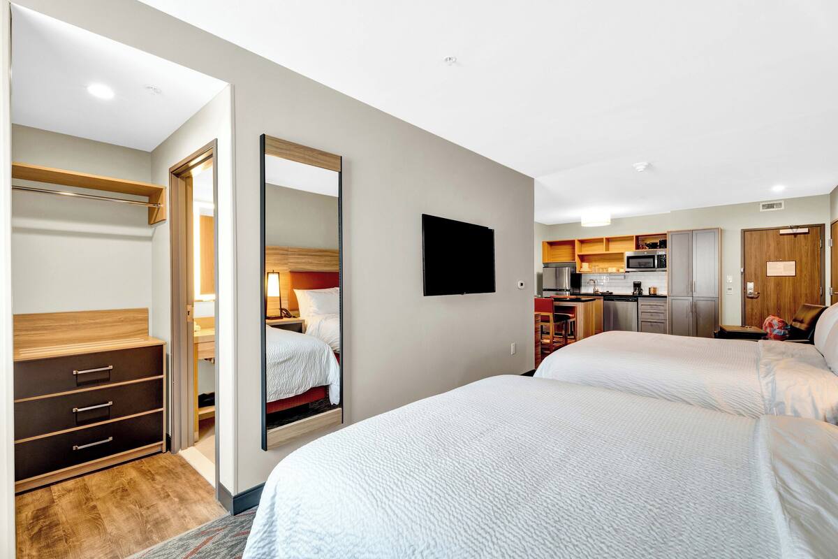 A hotel room with two beds, a wardrobe, a mirror, a television, and a kitchenette.