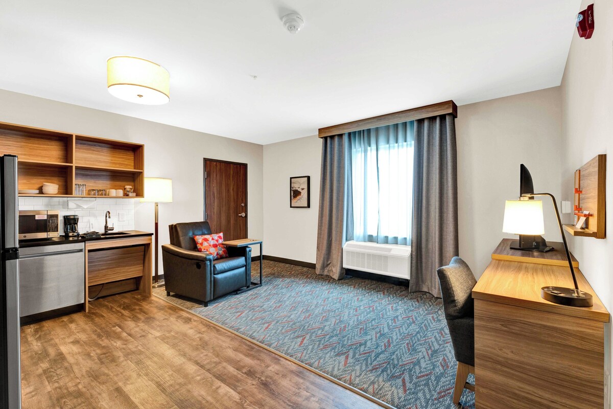 Une chambre d’hôtel dotée d’une kitchenette, d’un coin salon avec un fauteuil et un bureau, d’une grande fenêtre avec des rideaux et d’un tapis à motifs.