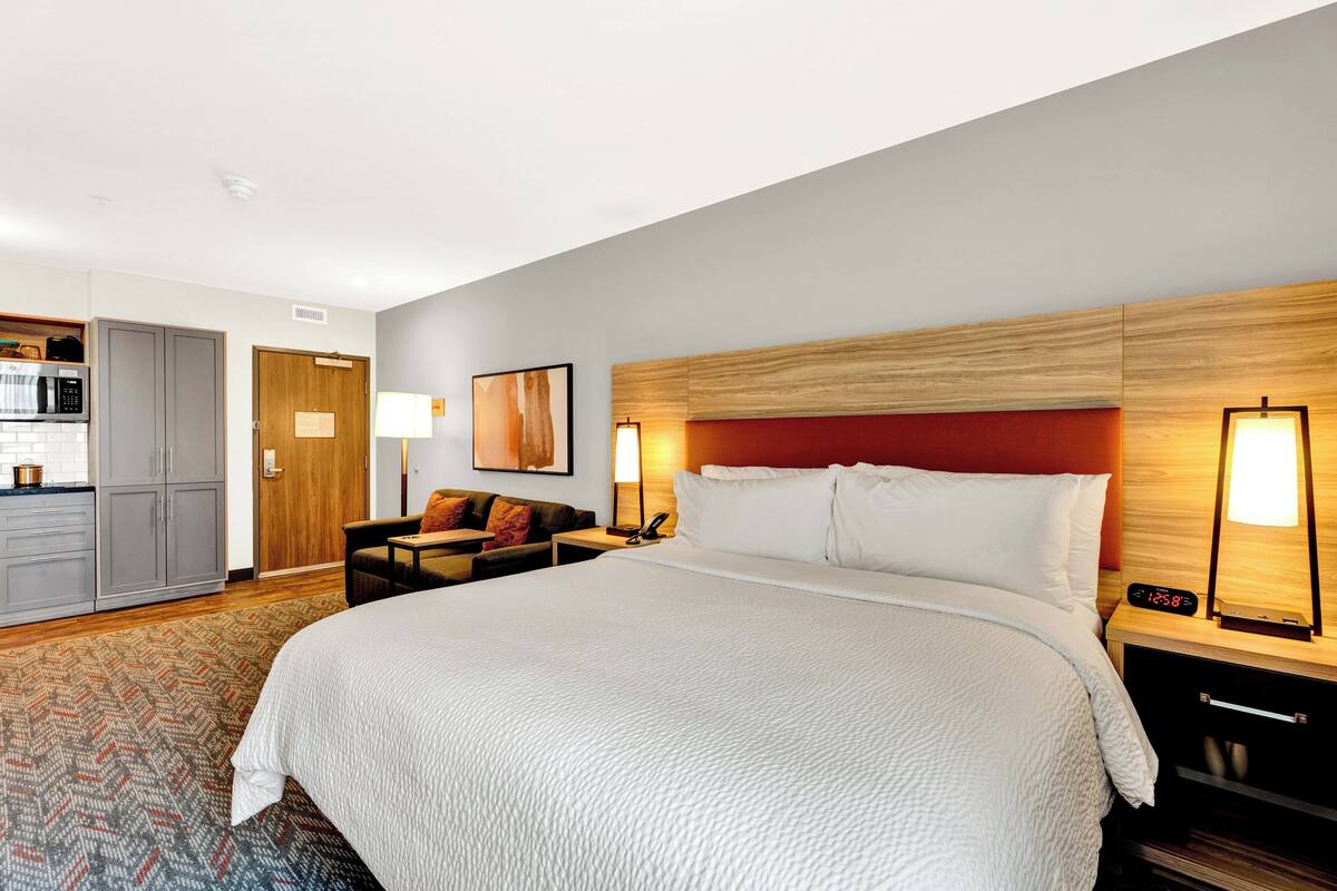 A hotel room with a large bed, a desk, a chair, a microwave, a refrigerator, and a door.
