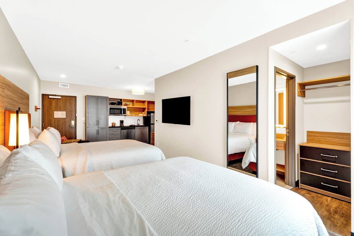 A hotel room with two beds, a kitchenette, a TV, a wardrobe, and a mirror.