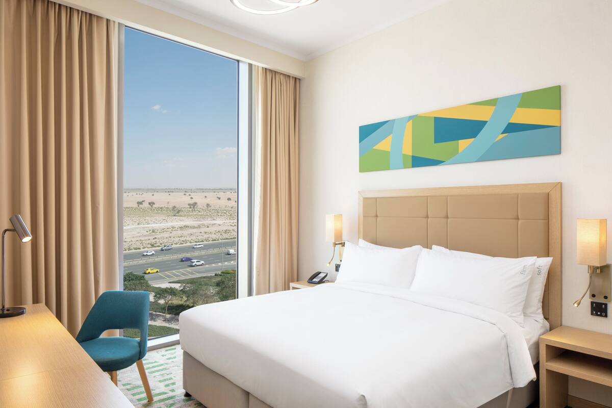 Hotelkamer met een groot bed, een bureau, een stoel en uitzicht op een woestijnlandschap door een groot raam.