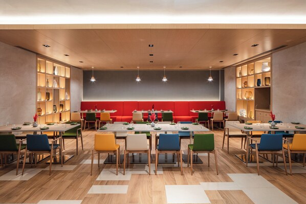 A modern dining area with multiple tables and chairs, a central red seating area, and built-in shelving with decorative items. - Holiday Inn Hotel & Suites Dubai Science Park by IHG (Dubai)