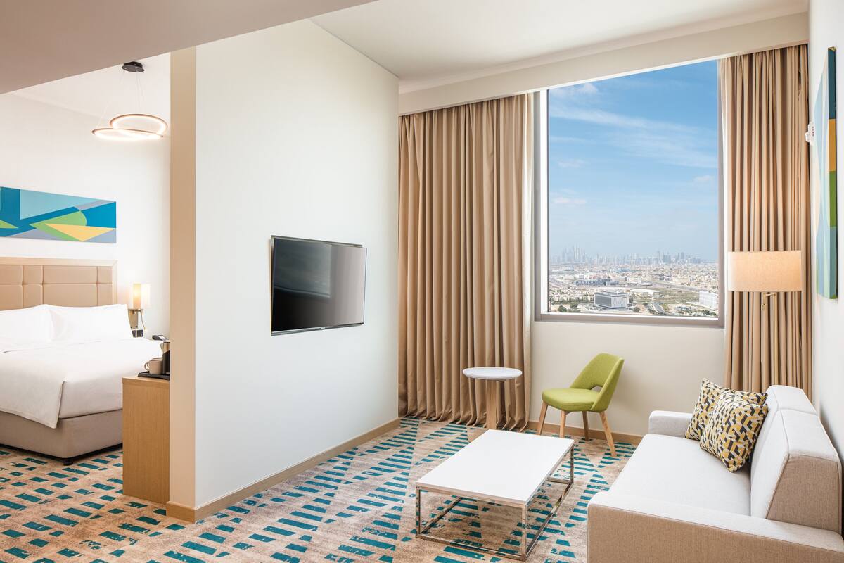 Een hotelkamer met een bed, een televisie, een bank, een stoel, een klein tafeltje en een groot raam met uitzicht over de stad.