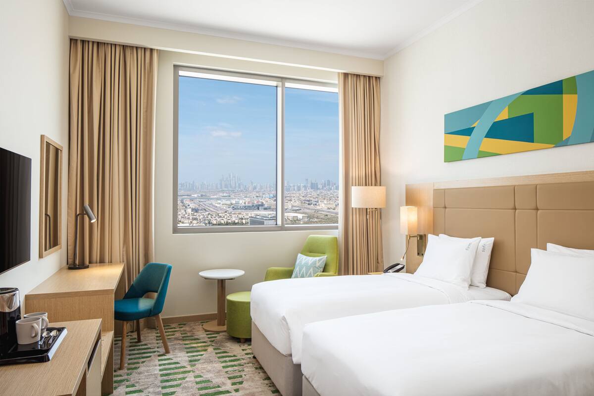 Een hotelkamer met twee bedden, een bureau met koffiezetapparaat, een televisie, uitzicht op de stad en een schilderij aan de muur.